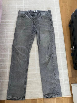 Grå raka jeans med slitning - Säljer ett par grå jeans med rak passform och klassisk femficksdesign. Jeansen har en diskret slitning på höger ben och är tillverkade i denim. Perfekta för en avslappnad och trendig look.