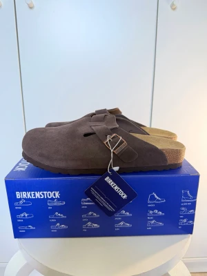 Birkenstock Boston Mocha tofflor - Säljer ett par Birkenstock Boston tofflor i färgen mocha. De är tillverkade i mjukt mockamaterial med en justerbar rem och metallspänne. Sulan är ergonomiskt utformad med kork och gummi för extra komfort. Perfekta för dig som vill ha både stil och bekvämlighet.