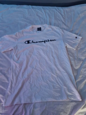 Vit Champion t-shirt med logga - Säljer en klassisk vit t-shirt från Champion i storlek medium. T-shirten har korta ärmar och stor svart Champion-logga tryckt över bröstet. Tillverkad i mjuk bomull och har en avslappnad passform. Perfekt till jeans eller shorts.