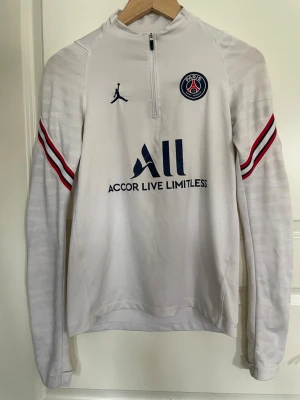 Vit PSG träningströja Jordan - Snygg vit långärmad träningströja från PSG och Jordan med halv dragkedja. Tröjan har blå och röda detaljer på ärmarna, PSG-logga på bröstet och tryck fram och bak. Perfekt för fotbollsträning eller chill. Materialet är troligtvis polyester.