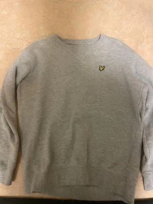 Grå sweatshirt från Lyle & Scott - Klassisk grå sweatshirt från Lyle & Scott med rund halsringning och ribbade muddar. Tröjan har den ikoniska gula örnloggan broderad på bröstet. Perfekt basic-plagg i mjukt bomullsmaterial som funkar till det mesta.