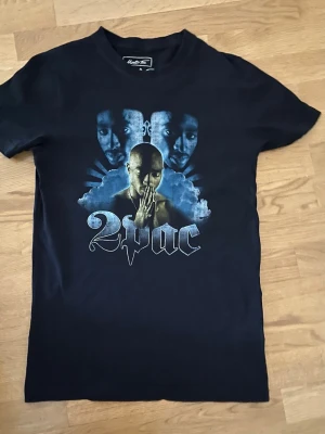 Svart 2Pac t-shirt från Mister Tee - Säljer en svart t-shirt från Mister Tee med stort 2Pac-tryck på framsidan i blåa och vita toner. T-shirten har klassisk passform och korta ärmar. Perfekt för dig som gillar streetwear och vill ha en ikonisk look.