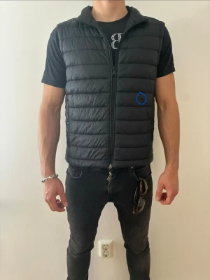 Boss väst - Snygg svart dunväst med horisontella sömmar och hel dragkedja framtill. Perfekt att slänga över en hoodie eller t-shirt för extra värme. Lätt och smidig att bära. Minimal defekt framtill, inringat i blått på första bilden, man ser knappt den.