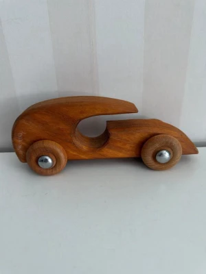 Träbil i retrodesign - Säljer en handgjord träbil med rundade former och naturlig träfärg. Bilen har fyra hjul med metallknoppar i mitten och ett öppet grepphål i karossen. Perfekt som dekoration eller leksak för den som gillar klassisk design.
