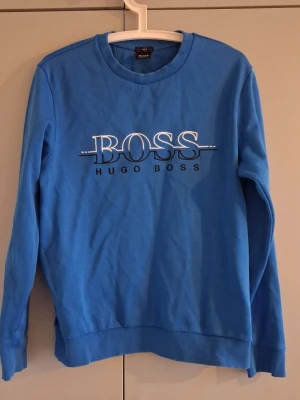 Blå sweatshirt från Hugo Boss - Snygg blå sweatshirt från Hugo Boss med stort BOSS-tryck och logga på bröstet. Tröjan har rund halsringning, långa ärmar och ribbade muddar vid ärmslut och nederkant. Perfekt för en avslappnad och stilren look.