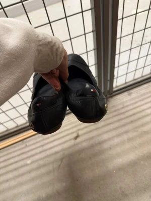 Svarta ballerinas från Tommy Hilfiger - Säljer ett par klassiska svarta ballerinas från Tommy Hilfiger i mjukt läder. Skorna har rund tå, diskret logotyp på hälen och en liten metallknapp på sidan. Perfekta för dig som gillar stilrena och bekväma skor.