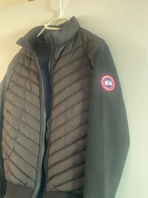 Svart pufferjacka från Canada Goose - Snygg svart pufferjacka från Canada Goose med quiltad front och mjuka stickade ärmar. Jackan har hög krage, dragkedja och den klassiska logotypen på vänster ärm. Perfekt för kalla dagar och riktigt stilren look.