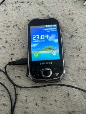 Samsung svart pekskärmsmobil - Säljer en svart Samsung mobiltelefon med pekskärm och fysiska knappar under skärmen. Telefonen har rundade kanter och en klassisk design med silverfärgade detaljer runt knapparna. Perfekt för dig som gillar retro eller behöver en enkel mobil.
