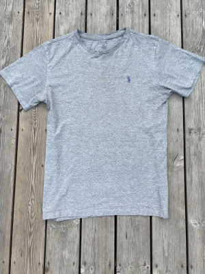 Grå t-shirt från Polo Ralph Lauren - Klassisk grå t-shirt från Polo Ralph Lauren med rund hals och korta ärmar. Diskret broderad logga i blått på bröstet. Tillverkad i mjuk bomull för en bekväm känsla och enkel stil.
