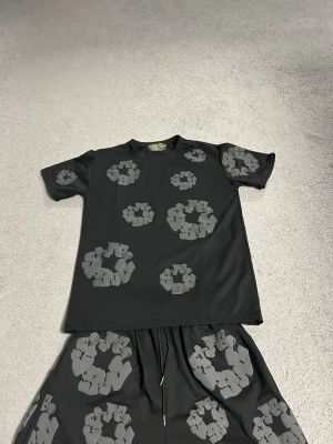 Svart set med gråa mönster - Säljer ett svart set med t-shirt och shorts, dekorerat med stora gråa blommönster över hela plaggen. T-shirten har rund hals och korta ärmar, och shortsen har resår i midjan. Perfekt för dig som gillar unika och trendiga prints.