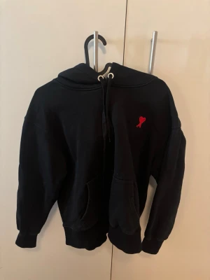 Svart hoodie med röd Ami-de-Coeur - Säljer en svart hoodie från AMI Paris med deras ikoniska röda Ami-de-Coeur broderat på bröstet. Hoodien har huva med dragsko, känguruficka och ribbade muddar. Tillverkad i mjuk bomull för skön känsla och avslappnad passform. Lite stor i storlek.