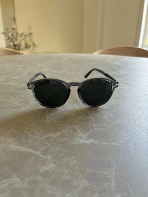 Svarta runda solglasögon från Tom Ford - Snygga svarta solglasögon från Tom Ford med rundad form och mörka glas. Bågarna är i plast med en transparent detalj vid näsan och klassisk design som passar till alla outfits. Perfekta för soliga dagar och ger en cool vibe.