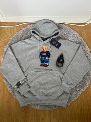Grå hoodie Polo Bear  - Säljer en grå hoodie med den ikoniska Polo Bear-figuren på bröstet. Hoodien har huva med dragsko och stor magficka. Materialet är mjukt och skönt bomullstyg. Perfekt för dig som gillar streetwear och klassisk stil.