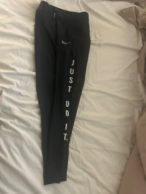 Svarta Nike tights med tryck - Svarta sporttights från Nike med vitt 'JUST DO IT.'-tryck längs ena benet och klassisk Nike-logga framtill. Tight passform och elastiskt material som passar perfekt till träning eller löpning. Oanvända! 