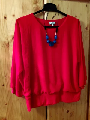 Röd chiffongblus,M - Röd blus med 3/4  ärmar och resår i ärmslut och nederkant från Manda Collection.  Blusen har en rund halsringning. Halsbandet ingår ej. Blusen är tillverkad av chiffong och har en mjukt material undertill.  Inte genomskinlig.  62% polyester, 33% bommul, 5% elastan. Storlek L.  Från armhålan till armhålan 57cm. Längd 60cm. Fint skick.