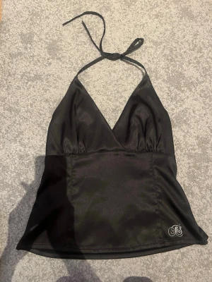 Svart halterneck blus i satin - Snygg svart halterneck blus i glansigt satinmaterial med djup v-ringning och knyt i nacken. Liten broderad logga nertill. Perfekt för att lyfta vilken outfit som helst.
