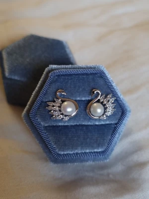 Eternal Love (silver 925 stift)  - Svanar är en symbol för kärlek, trohet och evighet – de representerar starka band och livslång samhörighet. Smycket är dekorerat med en sötvattenspärla, CZ-diamanter som reflekterar ljuset och ger en exklusiv lyster.                                    Material: Rhodium, Stift i 925 silver (stämplat), sötvattenspärlor, CZ-diamant Storlek: längd ca 1,5 cm Bredd ca 1,5 cm -Inga prisförslag, tack                                        - Varan packas noggrant och säkert 