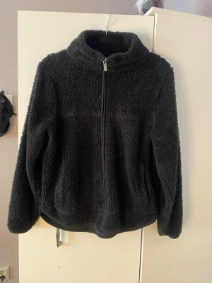 Svart teddyjacka från 157 - Mysig svart teddyjacka från 157 i storlek M. Jackan har dragkedja framtill, hög krage och är gjord i fluffigt teddy-material som håller dig varm. 