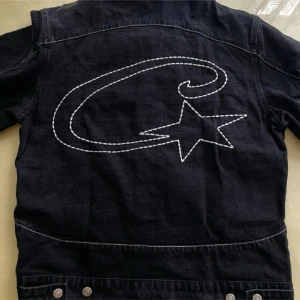 Svart jeansjacka med stjärndetalj - Cool svart jeansjacka med vit broderad stjärna och swirl på ryggen. Klassisk modell med krage, knappar framtill och bröstfickor. Perfekt för dig som vill sticka ut med en unik detalj på ryggen.