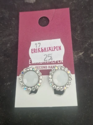 Silverfärgade clipsörhängen med stenar - Runda clipsörhängen i silverfärg med en stor vit sten i mitten, omgiven av små gnistrande stenar i olika färger. Klassisk och glittrig design som ger en elegant touch. Perfekta för dig som vill ha örhängen utan hål.