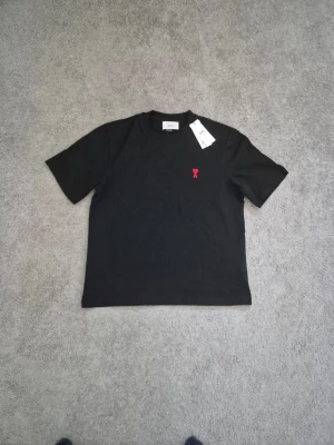 Ami - Svart t-shirt från Ami Paris med röd broderad logga på bröstet. Klassisk rund halsringning och korta ärmar. Tillverkad i mjuk bomull för en skön känsla. Perfekt för en clean och stilren look.