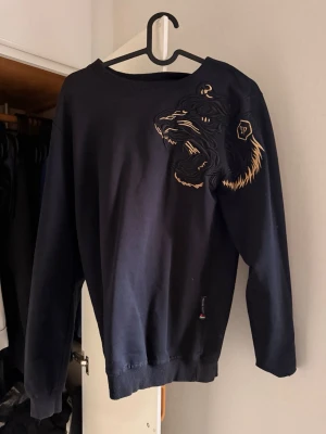 Svart sweatshirt med tigerbrodyr - Cool svart sweatshirt från Philipp Plein med stor broderad tiger i gult och svart på axeln. Tröjan har rund hals, långa ärmar och ribbade muddar. Perfekt för dig som gillar streetstyle och unika detaljer.