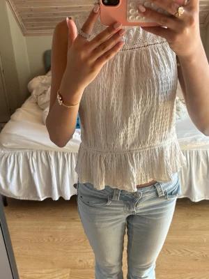 Vit ärmlös volangblus med spets - Säljer en söt vit ärmlös blus med volangkant och spetsdetaljer upptill. Blusen har en lätt skrynklig struktur och rund halsringning. Perfekt till jeans eller kjol för en fräsch och somrig look. 