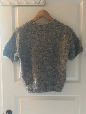 Grå stickad t-shirt i mohairmix - Mysig grå stickad t-shirt med korta ärmar och ribbstickad hals, ärmslut och nederkant. Tröjan har en fluffig struktur och är troligtvis stickad i en mjuk mohair- eller ullmix. Perfekt för lager på lager och en avslappnad look.