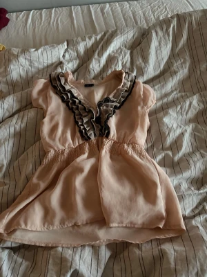 Ljusrosa blus med volangdetalj - Söt ljusrosa blus med korta ärmar och volangkant i svart och beige längs v-ringningen. Blusen har resår i midjan för en snygg passform och är tillverkad i ett tunt, lätt tyg som ger ett luftigt intryck. Köpte denna på plick men säljer då den var för stor!❤️