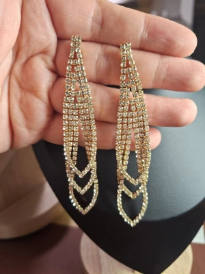 Långa guldiga strassörhängen - Säljer ett par långa, glittriga örhängen i guldton med flera rader av små strass-stenar. Örhängena har en elegant, droppformad design med geometriska detaljer som ger en lyxig känsla. Perfekta för dig som vill sticka ut och addera extra bling till din look.