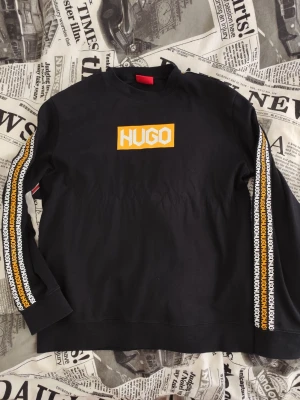Svart sweatshirt från Hugo Boss - Svart sweatshirt från Hugo Boss med stort gult och vitt HUGO-tryck på bröstet. Längs båda ärmarna löper ett band med upprepat HUGO-tryck i vitt och gult. Tröjan har rund halsringning och ribbade muddar. Perfekt för en streetwear-look.