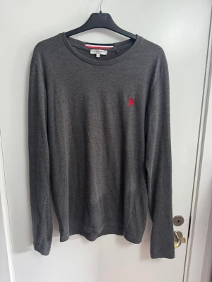 Mörkgrå tröja från U.S. Polo Assn - Mörkgrå långärmad tröja från U.S. Polo Assn med rund halsringning och röd broderad logga på bröstet. Tröjan har en enkel och stilren design, perfekt för dig som gillar klassiska plagg med en twist. Lite nopprig ses på bilder.