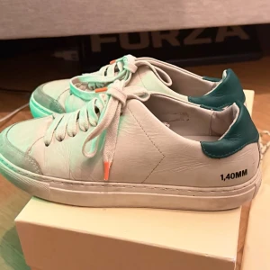 Axel Arigato sneakers - Snygga vita sneakers från Axel Arigato med gröna detaljer på hälen och texten '1,40MM' på sidan. Skorna har klassisk låg profil, vita snören med orange detalj och är tillverkade i läder. Perfekta för en stilren och trendig look.
