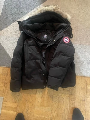 Svart dunjacka från Canada Goose - Säljer en svart dunjacka från Canada Goose med avtagbar huva och pälskant. Jackan har klassisk patch-logga på ärmen, dragkedja och knappar framtill samt stora fickor. Perfekt för kalla vinterdagar och riktigt varm tack vare dunfyllningen.