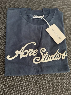 Blå T-Shirt från Acne Studios - Snygg blå t-shirt från Acne Studios med stort vitt logotyp-tryck framtill. Klassisk rund halsringning och korta ärmar. Tillverkad i mjuk bomull för en skön känsla. Perfekt för dig som gillar stilrena och trendiga plagg. NYPRIS: 3800kr