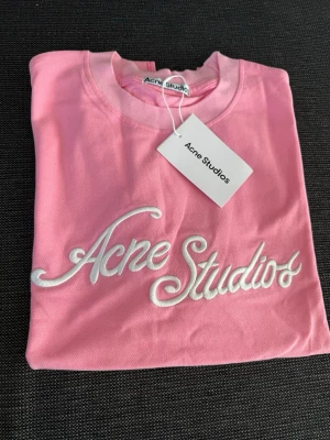 Rosa Acne Studios t-shirt  - Säljer en rosa t-shirt från Acne Studios med vit broderad logga framtill. T-shirten har klassisk passform, rund halsringning och är kortärmad. Tillverkad i mjuk bomull och perfekt för en avslappnad och trendig look. NYPRIS: 3800kr