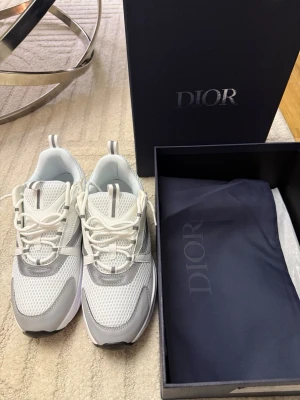 Dior b22 - Snygga sneakers från Dior i vitt och grått med mesh och läderdetaljer. Skorna har en modern och sportig design med vita snören och diskreta logodetaljer. Perfekta för dig som vill ha en exklusiv och trendig look.