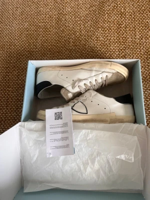 Vita Phillipe modell  - Stilrena vita sneakers med svart detalj på hälen och klassisk låg profil. Skorna har vita snören, perforerade sidor och en något tjockare sula. Tillverkade i läder för en clean look som passar till det mesta. Qr kod finns för checka om det är riktiga vilket de är. Priset är förhandlingsbart så kontakta bara😀