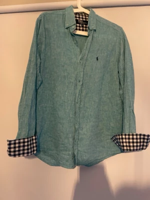 Ljusblå linneskjorta Polo Ralph Lauren - Snygg ljusblå skjorta i linne från Polo Ralph Lauren, slim fit. Skjortan har långa ärmar och kontrasterande rutigt tyg på insidan av manschetterna. Klassisk krage och liten broderad logga på bröstet.