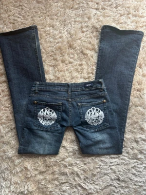 Mörkblå jeans med dödskallebrodyr - Säljer ett par mörkblå jeans med utsvängda ben och coola vita dödskallebroderier på bakfickorna. Jeansen har klassisk femficksmodell, kontrastsömmar och detaljer i mässing. Perfekta för dig som vill sticka ut med en edgy stil. Org pris- ca 1500