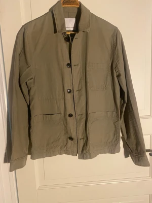 Beige overshirt från Samsøe Samsøe - Stilren beige overshirt från Samsøe Samsøe med raka linjer och tre utanpåliggande fickor framtill. Tillverkad i ett mjukt bomullstyg och har klassisk krage samt knäppning med svarta knappar. Perfekt lager på lager-plagg för en avslappnad look.