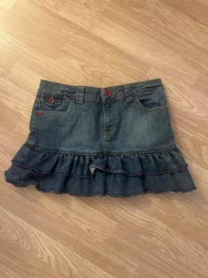 Jeanskjol med volang - Snygg jeanskjol i blå denim med dubbla volanglager nertill och röda knappdetaljer. Kjolen har både fram- och bakfickor samt hällor för skärp. 💗