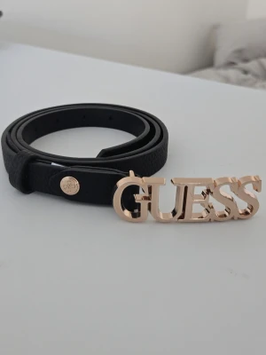 Svart bälte med guldspänne från Guess - Svart bälte från Guess med ett guldfärgat spänne med GUESS loggan. Bältet är smalt och har en stilren design! Har inte använt denna så många gånger, dock gjort egengjorda hål i den. Utöver det har den inga defekter. 