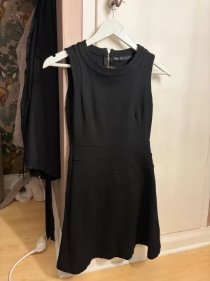Svart ärmlös klänning med dragkedja - Snygg svart ärmlös klänning från zara med rund halsringning och markerade sömmar i midjan. Klänningen har en guldig dragkedja längs ryggen och är i en kort modell med lätt utsvängd kjol. Perfekt för dig som gillar stilrena plagg.