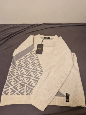 Gråvit stickad Fendi-tröja - Snygg stickad tröja från Fendi i gråvitt med ikoniskt FF-mönster på ena sidan och ärmen. Tröjan har rund halsringning, ribbade muddar och är tillverkad i mjukt material. Perfekt för dig som gillar exklusiva märken och stilren design.