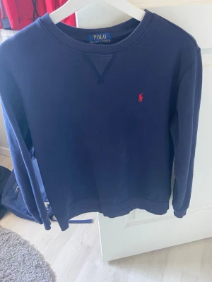 Mörkblå sweatshirt från Polo Ralph Lauren - Mörkblå sweatshirt från Polo Ralph Lauren med klassisk rund halsringning och röd broderad logga på bröstet. Tröjan har långa ärmar och är tillverkad i mjukt bomullsmaterial. Perfekt för en avslappnad och stilren look.