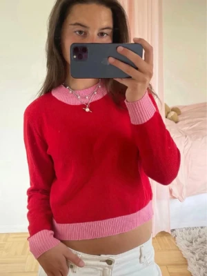 Röd stickad tröja med rosa detaljer - Snygg röd stickad tröja med kontrasterande rosa ribbade muddar, krage och nederkant. Tröjan har en klassisk rund halsringning och är figurnära i modellen. Perfekt för dig som vill ha en färgglad och trendig look.