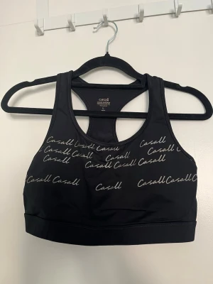 Svart sport-bh från Casall - Sport-bh från Casall i svart med vitt logotryck över hela framsidan. Modellen har brottarrygg och bred resår nedtill för extra stöd.