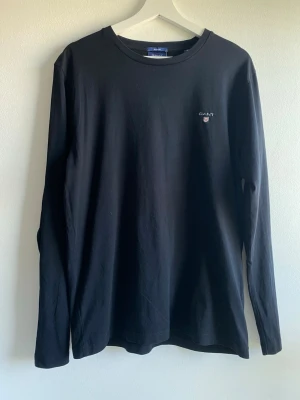 Svart långärmad tröja från GANT - En svart långärmad tröja från GANT med rund halsringning och liten broderad logga på bröstet. Modellen är regular fit och tillverkad i mjuk bomull, perfekt för en clean och stilren look.
