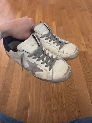 Golden Goose vita sneakers med grå stjärna - Snygga vita sneakers från Golden Goose med grå stjärna på sidan, grå skosnören och mörkblå häl. Skorna har en klassisk låg modell och är tillverkade i läder med slitna detaljer för en cool, avslappnad look.